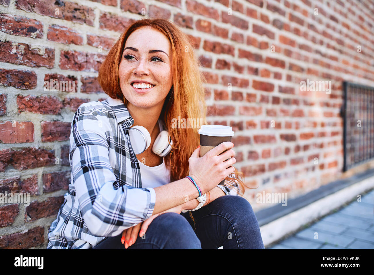 Giovane donna seduta contro un muro di mattoni holding tazza di caffè e guardando lontano Foto Stock