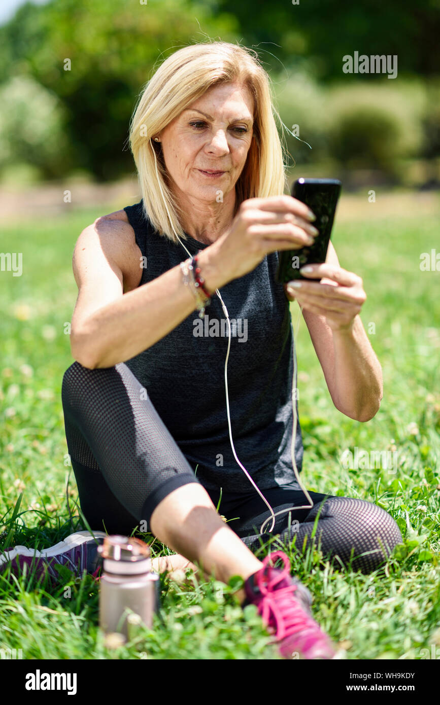 Donna matura, utilizzando il suo smartphone, appoggiata in erba di un parco dopo esercizio Foto Stock