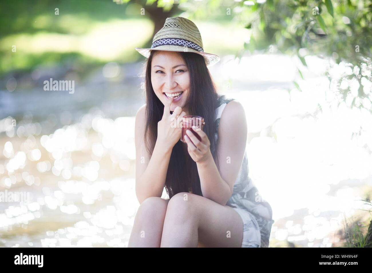 Sorridente giovane donna che indossa un cappello di paglia, seduto in riva al fiume e roditura Foto Stock