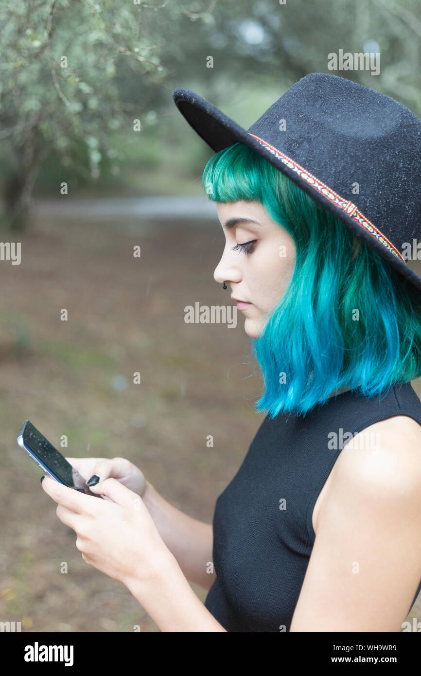 Profilo di giovane donna con tinti di blu e verde capelli utilizzando smartphone sul giorno di pioggia Foto Stock