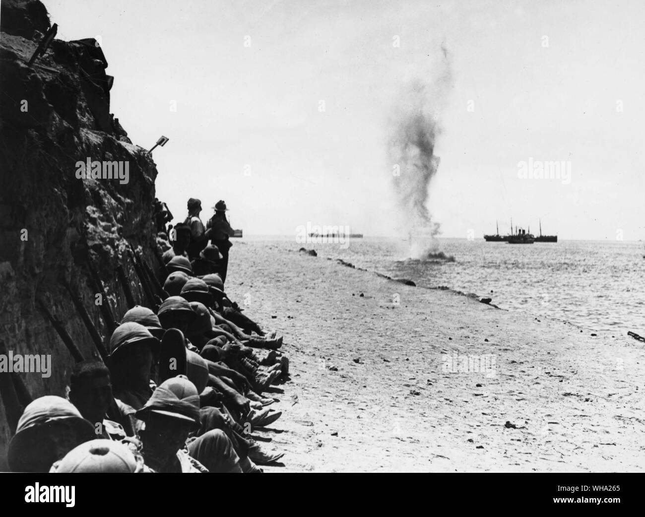 WW2: un guscio da Annie Asiatico, scoppio in mare. Gli uomini in appoggio sulla strada della spiaggia di Capo Helles al burrone burrone, sotto riparo della scogliera. Truppe alleate guarda su dalla spiaggia. Foto Stock