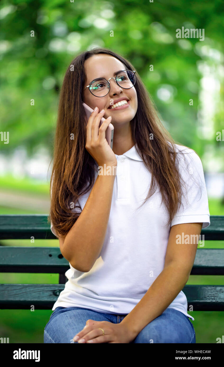 Ragazza giovane e carina con gli occhiali avente un telefono talk su una panchina nel parco Foto Stock