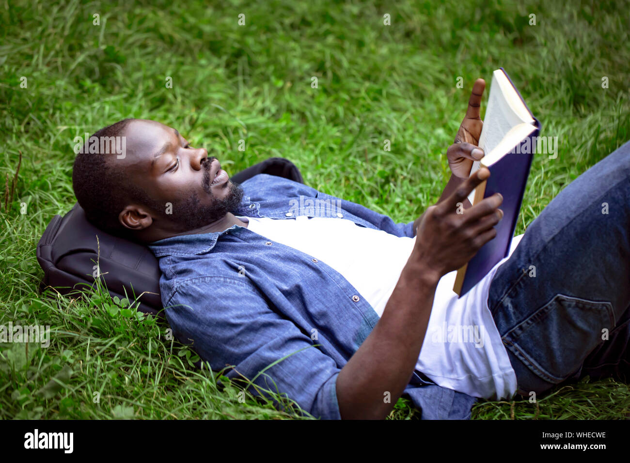 Concentrato maschio nero studente la lettura di un libro di testo sdraiati su un prato del campus Foto Stock