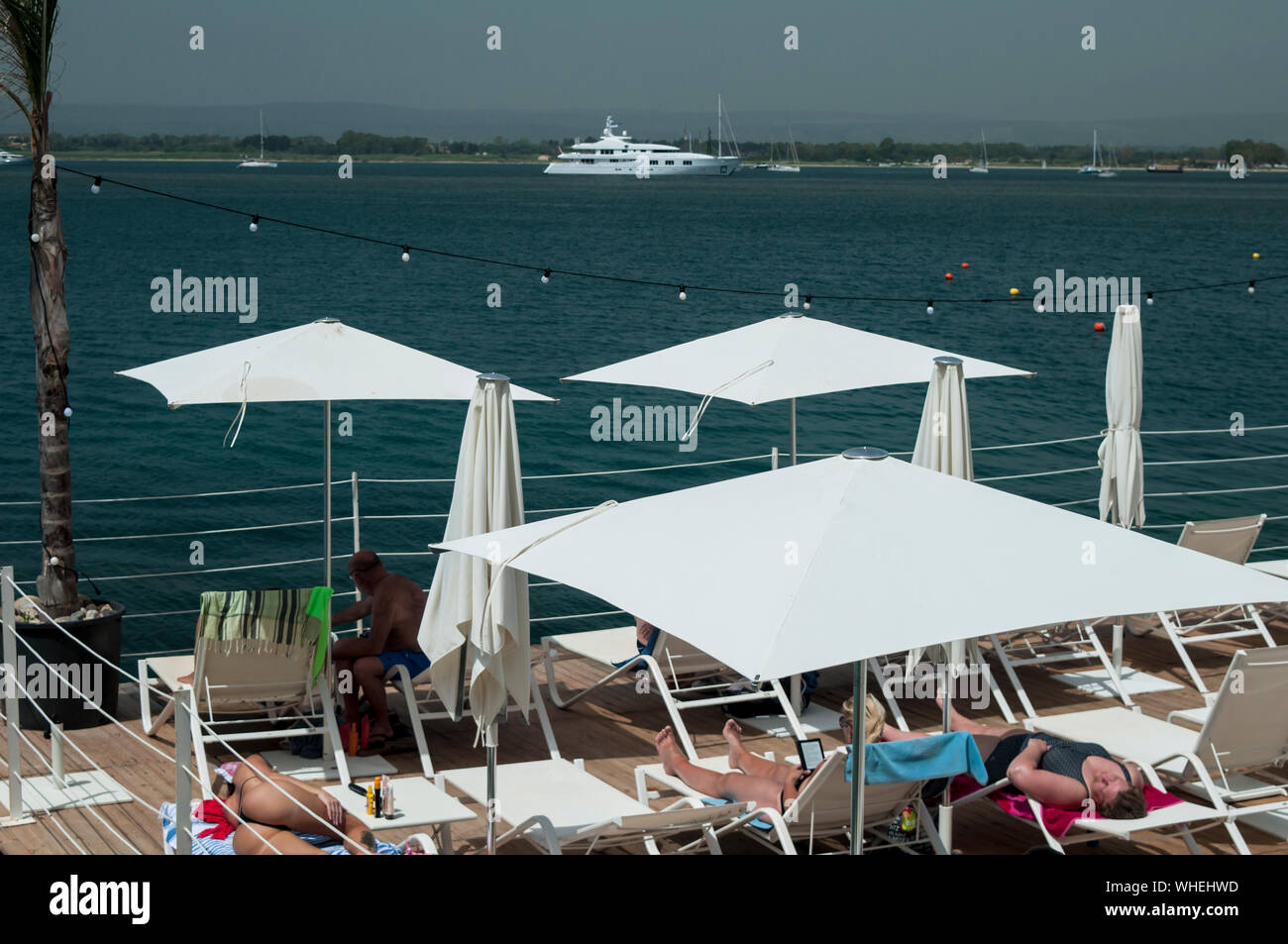 Ombrelloni bianchi e blu oceano offrono un ambiente perfetto per godersi un pomeriggio d'estate sull'isola di Ortigia, Siracusa, Sicilia. Uno yacht di lusso i Foto Stock