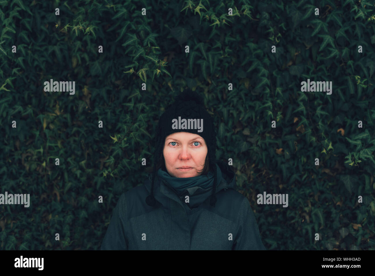 Grave donna in abiti invernali contro la parete di edera om fredda giornata invernale, guardando triste e preoccupato Foto Stock