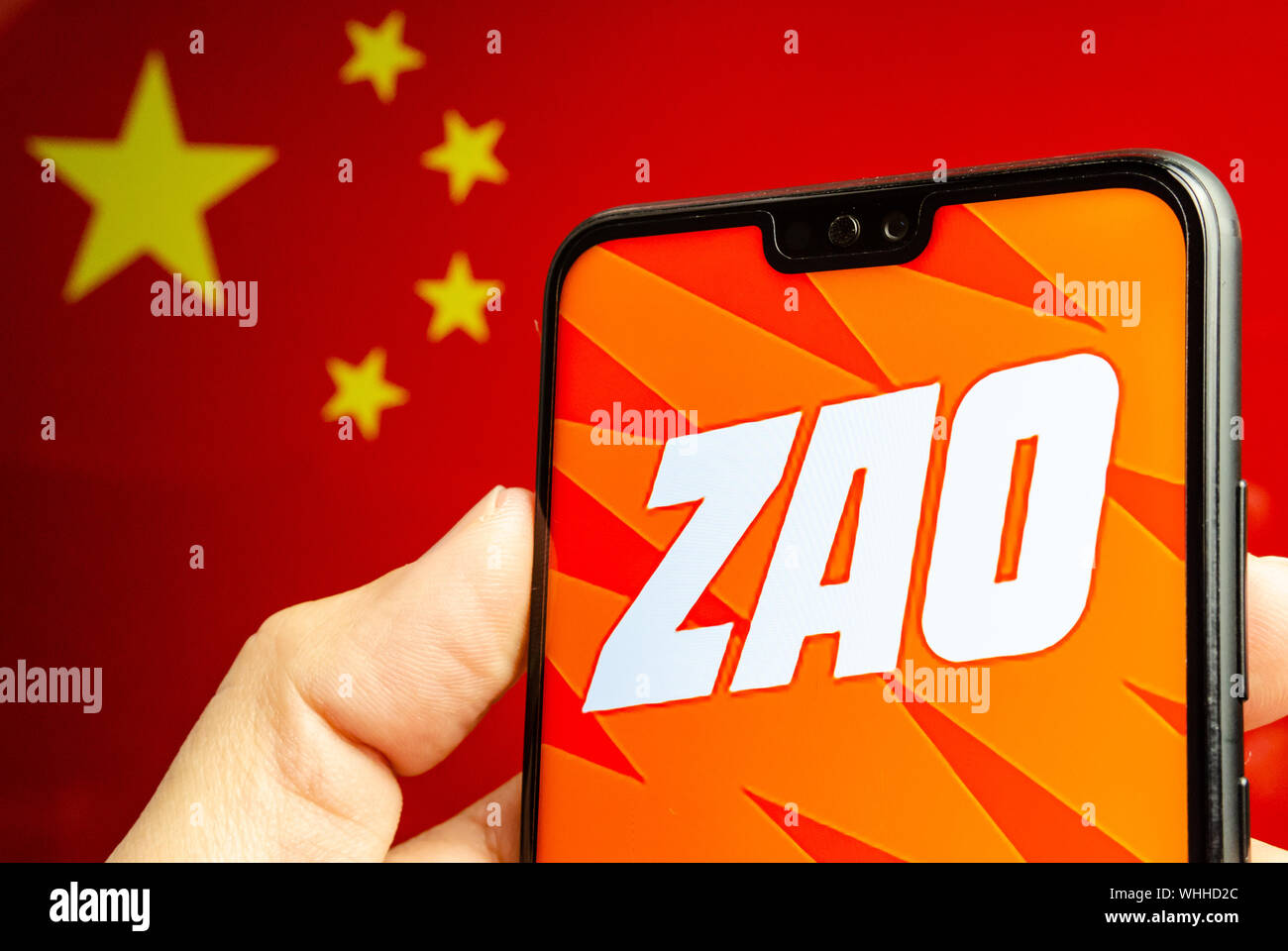 Mano trattiene un smartphone con ZAO app logo. La bandiera sfocata della Cina in background. Zao è attualmente un numero una divertente app in Cina. Foto Stock