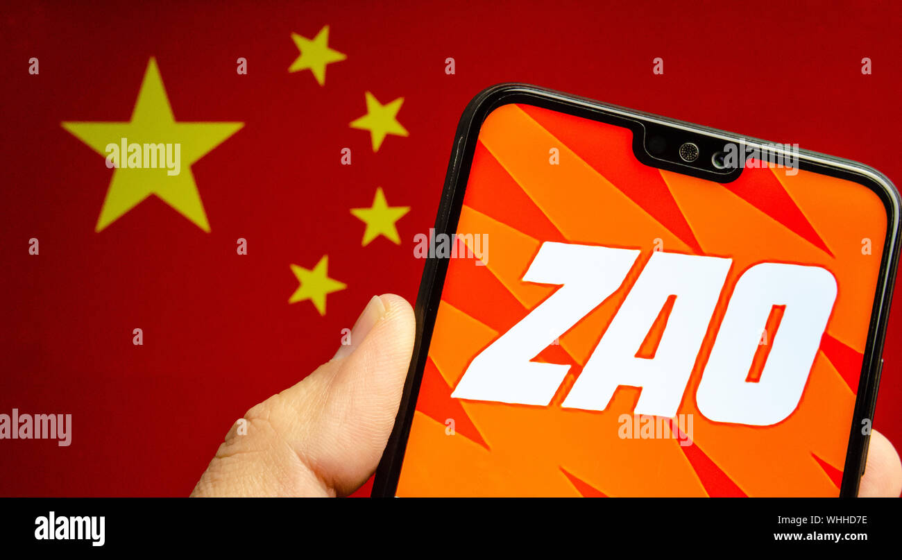 Mano trattiene un smartphone con ZAO app logo. La bandiera sfocata della Cina in background. Zao è attualmente un numero una divertente app in Cina. Foto Stock