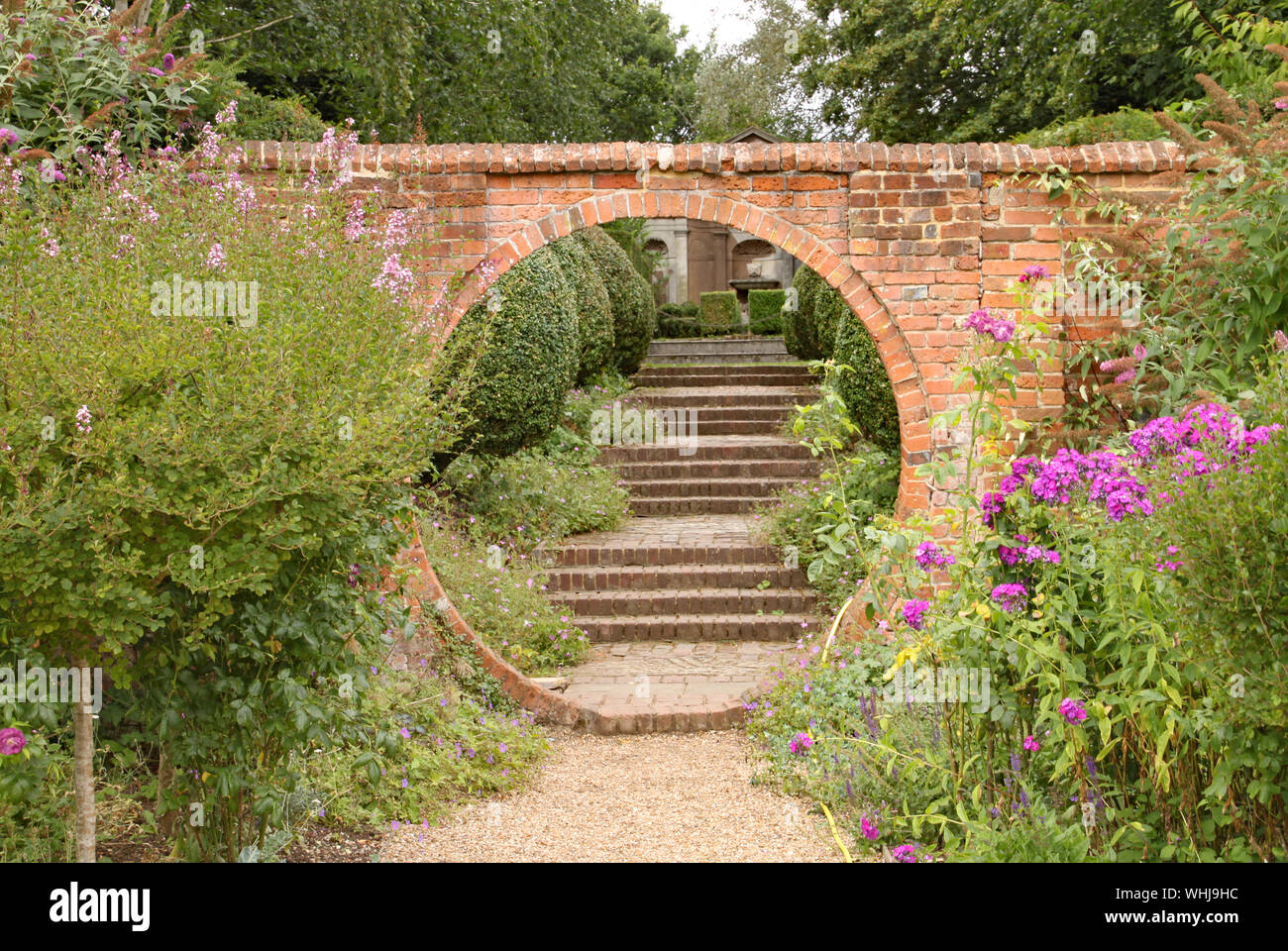 Un sentiero di ghiaia passa attraverso un arco circolare in un vecchio muro di mattoni, che conduce ad alcuni mattoni vecchi passi in un paese di lingua inglese giardino Foto Stock