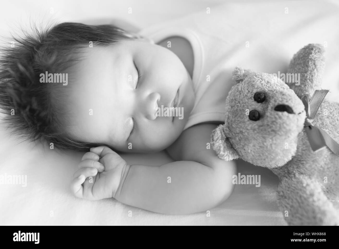 Adorable baby, pacificamente addormentato nel presepe accanto a un orsacchiotto di peluche su un fresco pomeriggio in bianco e nero. Foto Stock