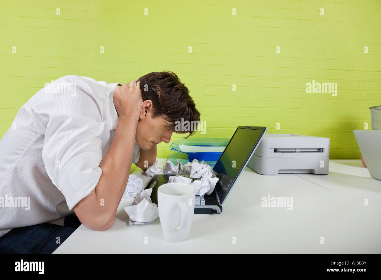 Vista laterale del frustrato giovane imprenditore con palline di carta e il laptop sulla scrivania Foto Stock