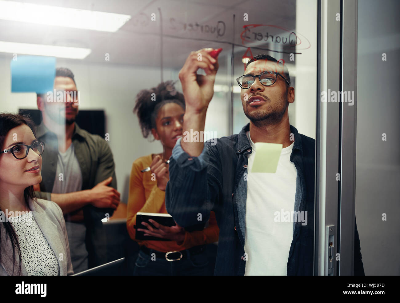 Collega di lavoro guardando imprenditore iscritto sulla parete di vetro con marcatore in ufficio Foto Stock