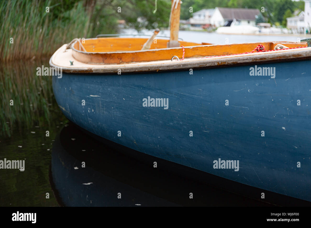 Chiudere la vista di una piccola barca a vela con scafo blu galleggiante su una via navigabile con case in background. Foto Stock
