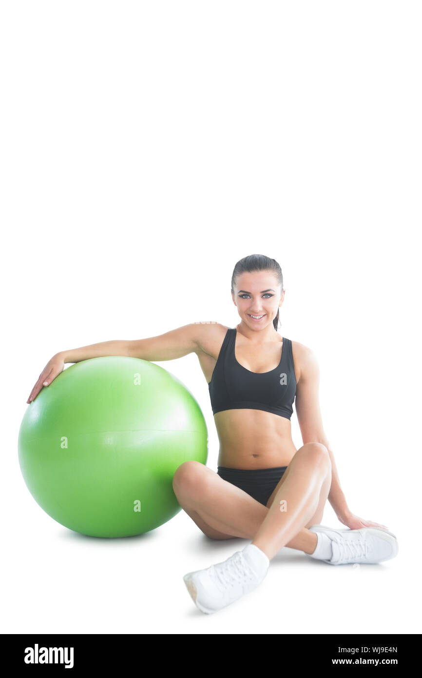 Vista frontale di attraenti brunette donna in posa seduta accanto a una palla ginnica su sfondo bianco Foto Stock