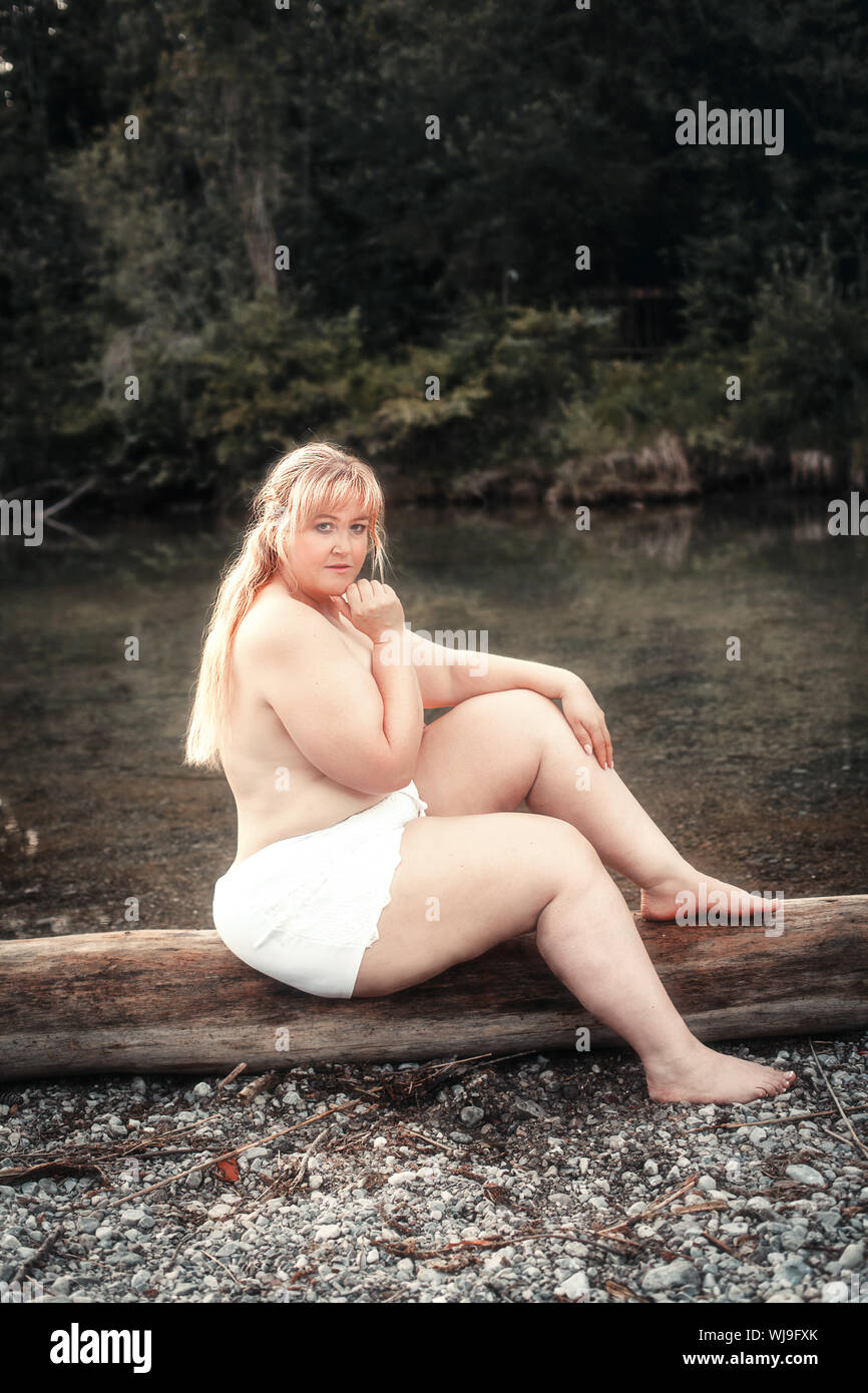 Giovane donna curva siede scarsamente rivestita su un lago su un tronco di albero Foto Stock