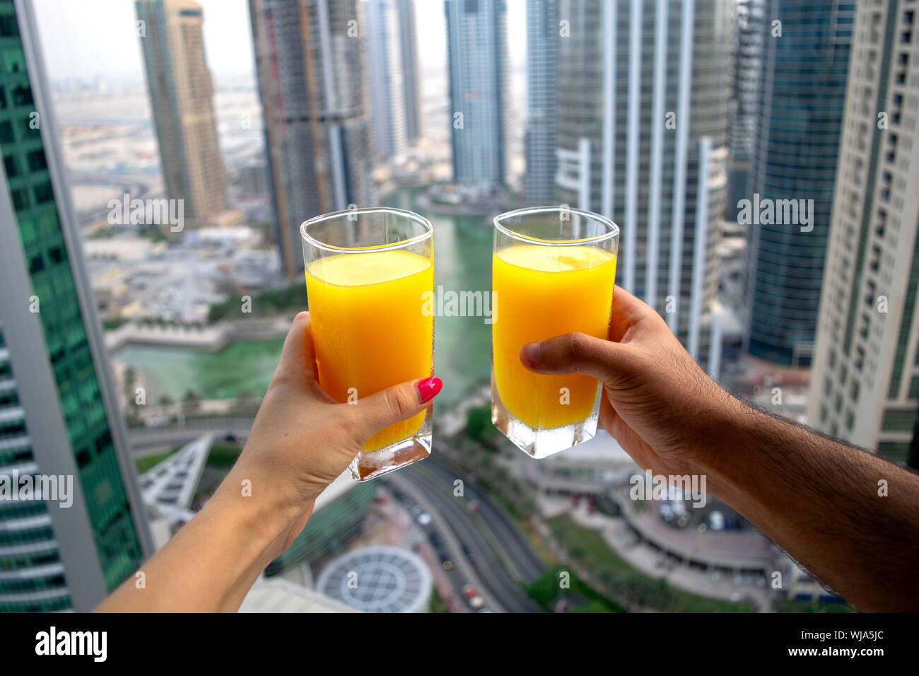 Maschi e femmine di tenere le mani di bicchieri di freddo fresco succo di arancia con città grattacieli in vista della città moderna di Dubai. Due bicchieri di succo di arancia sul cityscap Foto Stock