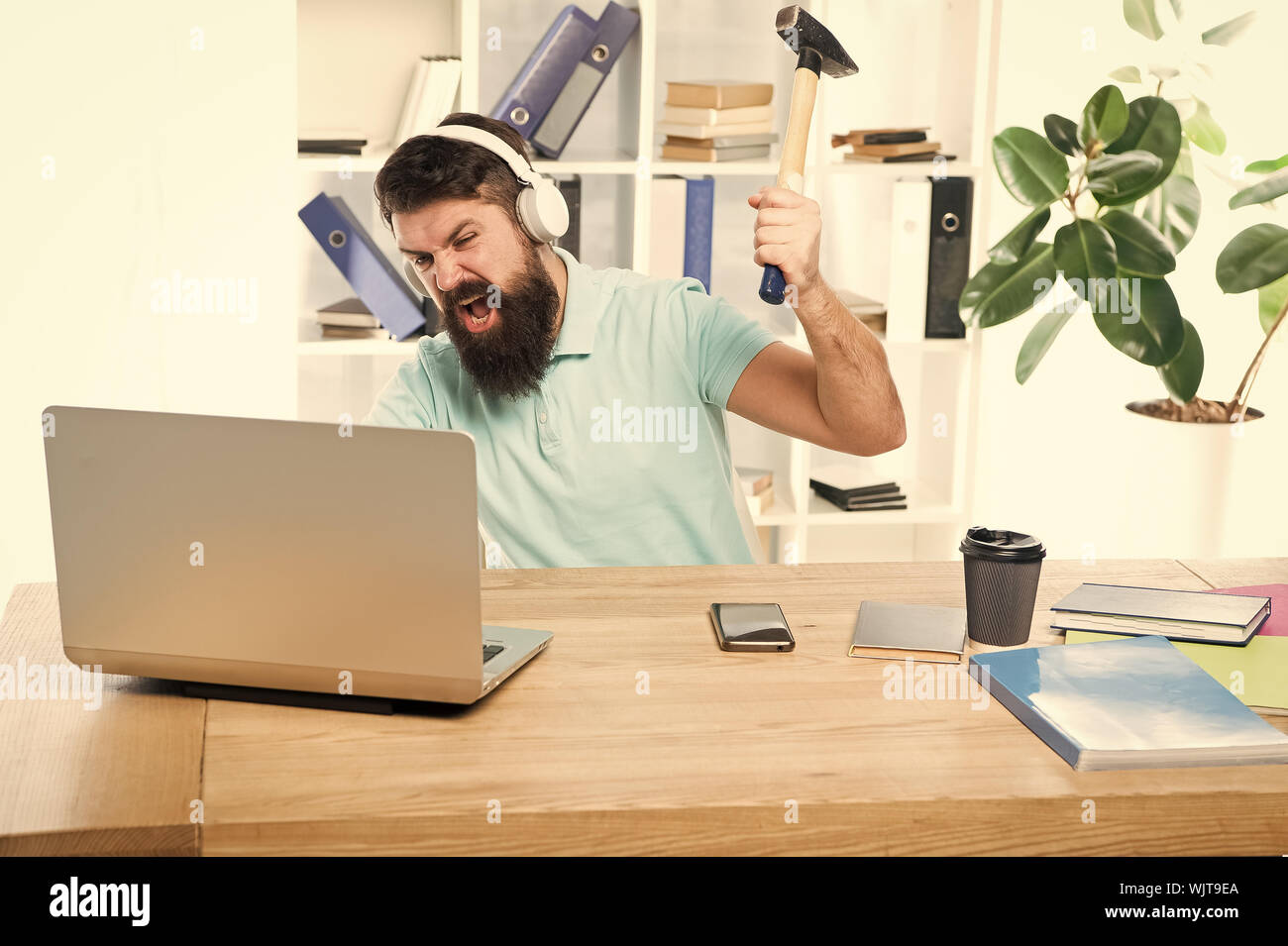 Uomo Barbuto guy ufficio cuffie swing martello sul computer. Connessione a internet lenta. Software obsoleto. Computer lag. Ragioni per computer in ritardo. Come fissare il lento sistema di ritardo. Odio di ufficio di routine. Foto Stock