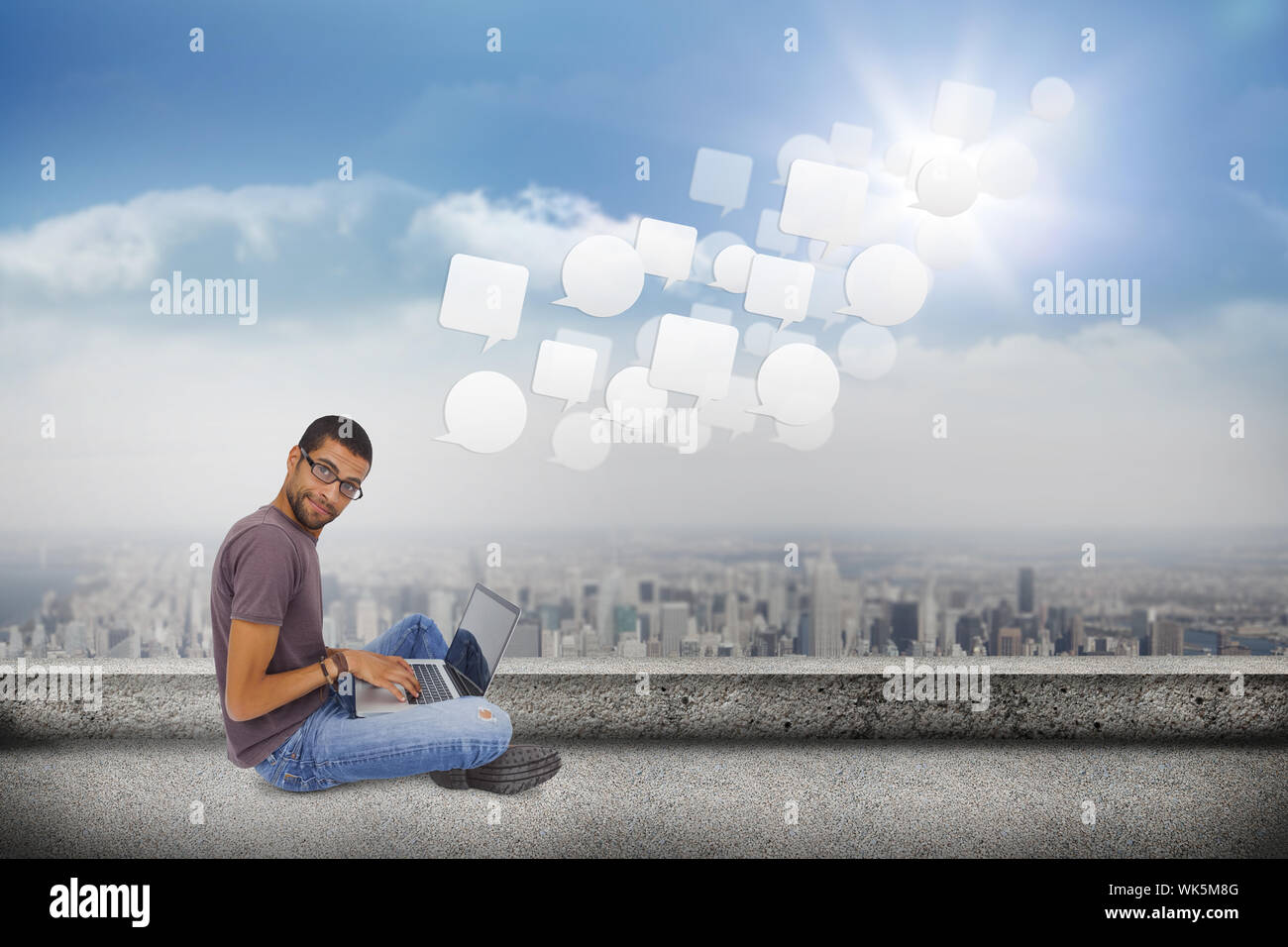 Uomo con gli occhiali seduto sul pavimento con il laptop e guardando la telecamera contro il balcone che si affaccia sulla città Foto Stock