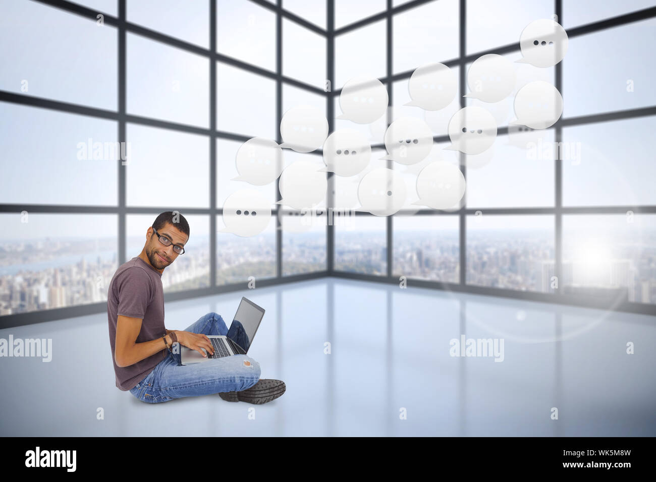 Uomo con gli occhiali seduto sul pavimento con il laptop e guardando la telecamera contro la camera con grandi finestre che mostra città Foto Stock