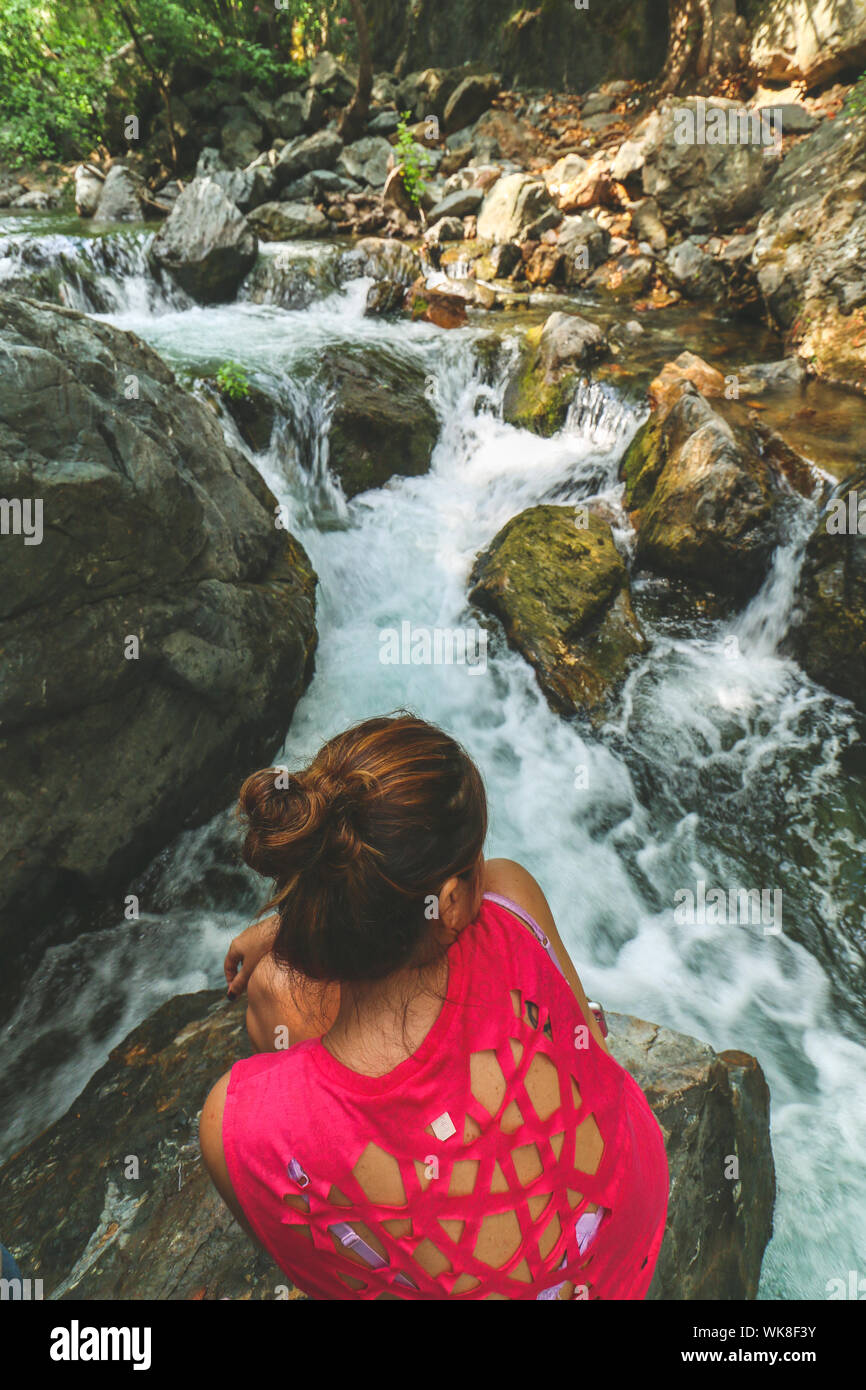 Giovane donna caucasica, indossa una maglietta rossa con forma netta tagli a forbice , è seduta su una grande roccia e guardando lo streaming di acqua di un fiume. Foto Stock