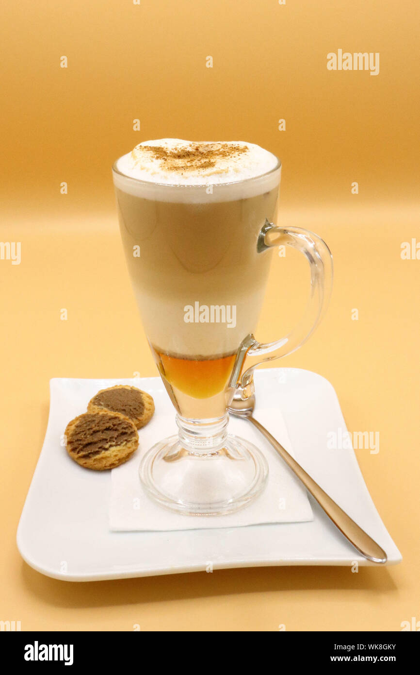 Tazza in vetro di latte macchiato a strati con schiuma di latte e cannella, servita con biscotti e cucchiaio su un piatto bianco, isolato su sfondo arancione Foto Stock
