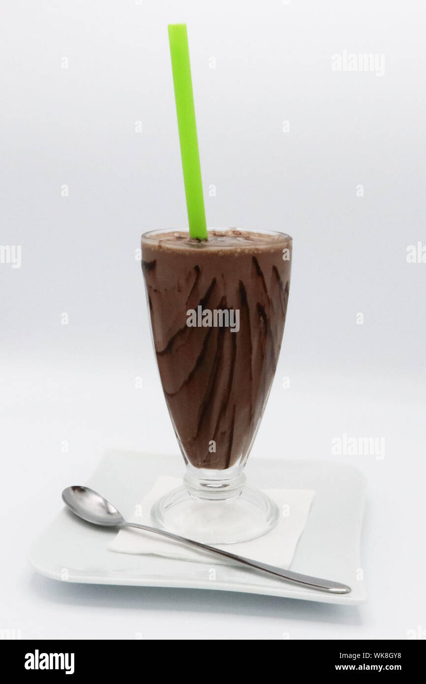 Rinfrescante frullato di cioccolato servito in un bicchiere alto con gocciolatoio di cioccolato, paglia verde e un cucchiaio su un piatto bianco. Foto Stock