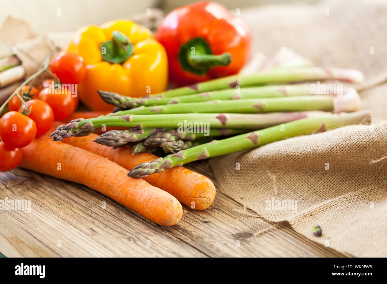 Ortaggi freschi in una cucina di paese con asparagi verdi spears,, pomodori ciliegia sulla vite, dolci peperoni e carote giacente un hessian waiti Foto Stock