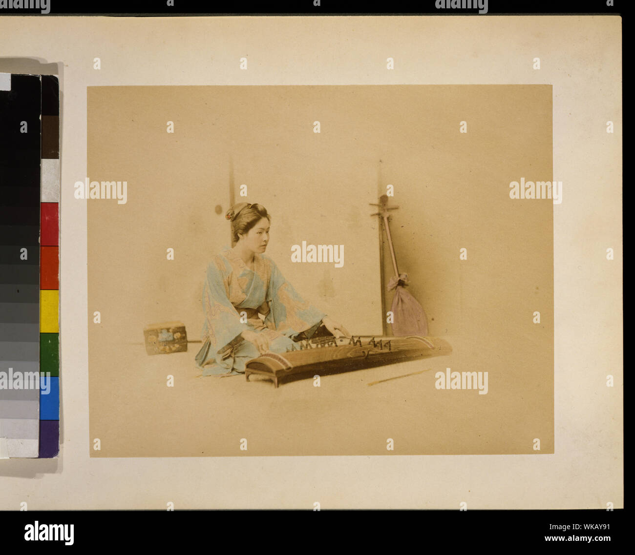 Donna Giapponese a piena lunghezza, seduto, rivolto verso destra, la riproduzione di un koto; un shamisen appoggia contro una parete in background Foto Stock