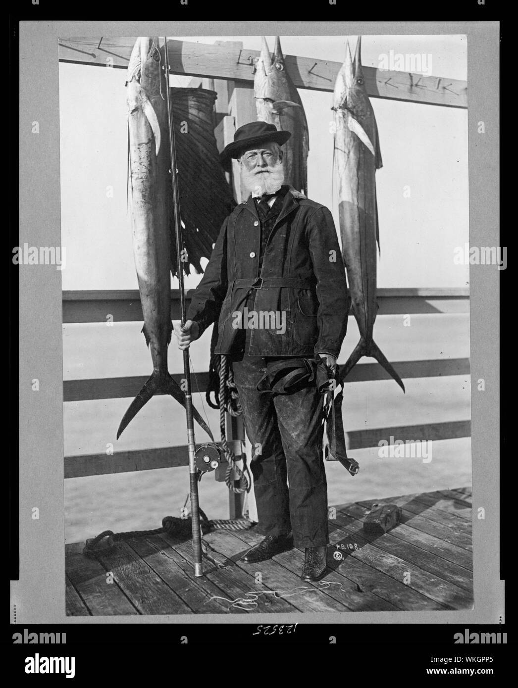 John D. Crimmins, ritratto a figura intera, in piedi sul molo, tenendo la canna da pesca e Mulinello, rivolto verso la parte anteriore, con tre pesci di mare appeso sul parapetto Foto Stock
