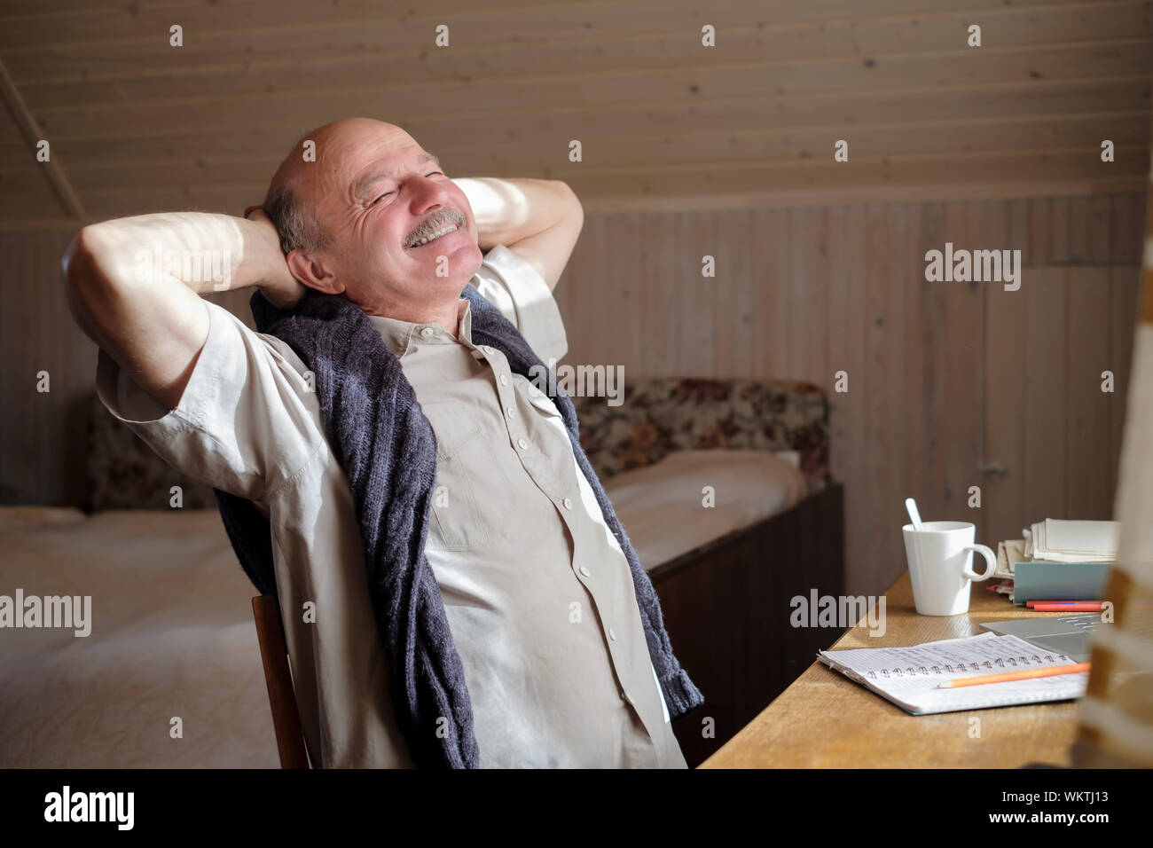 Rilassata senior uomo ispanico tenendo le mani dietro la testa e sorridente seduto al tavolo. Buon giorno sognatore concept Foto Stock