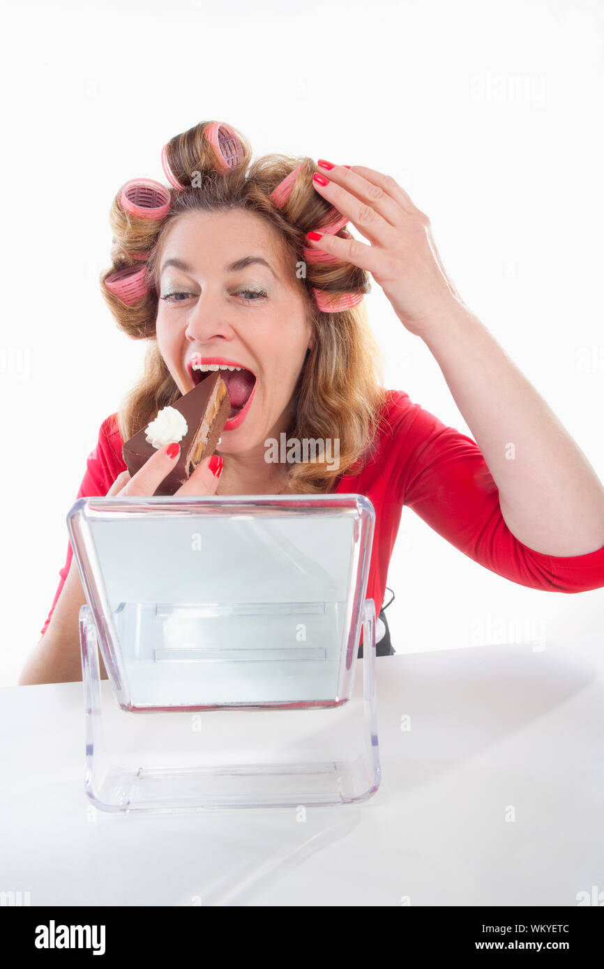 Donna di mezza età con i capelli rulli di mangiare la torta guardando lo specchio Foto Stock