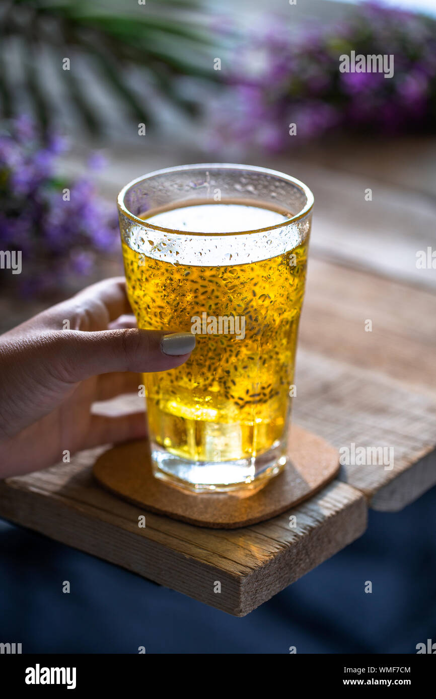 Tenere a mano un bicchiere di bevanda a base di semi di chia dorati su un tavolo di legno con fiori viola sullo sfondo. Foto Stock