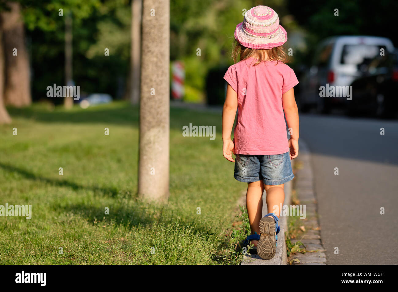 Vista posteriore di una ragazza di bambino in estate abbigliamento camminare da solo su un marciapiede tra un prato e una strada nella luce della sera di un giorno di estate Foto Stock