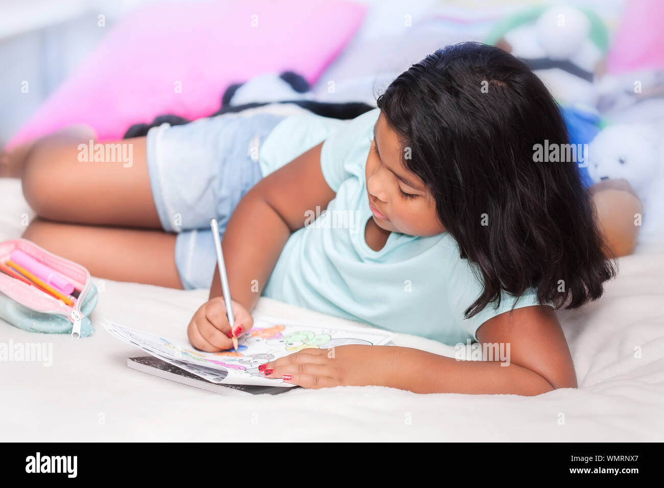 Una bimba di 8 anni, indossare pantaloncini denim facendo il suo dovere di matematica mentre posa nel suo letto comodo con animali impagliati in background. Foto Stock