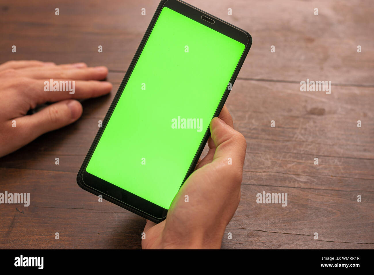 La mano della persona caucasica tenendo un nero smartphone non identificato con schermo verde su un tavolo di legno sfondo Foto Stock