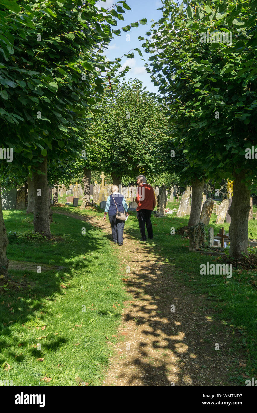 L uomo e la donna a piedi attraverso un paese di lingua inglese sagrato, il percorso illuminato da pezzata sole estivo; Long Buckby, Regno Unito Foto Stock