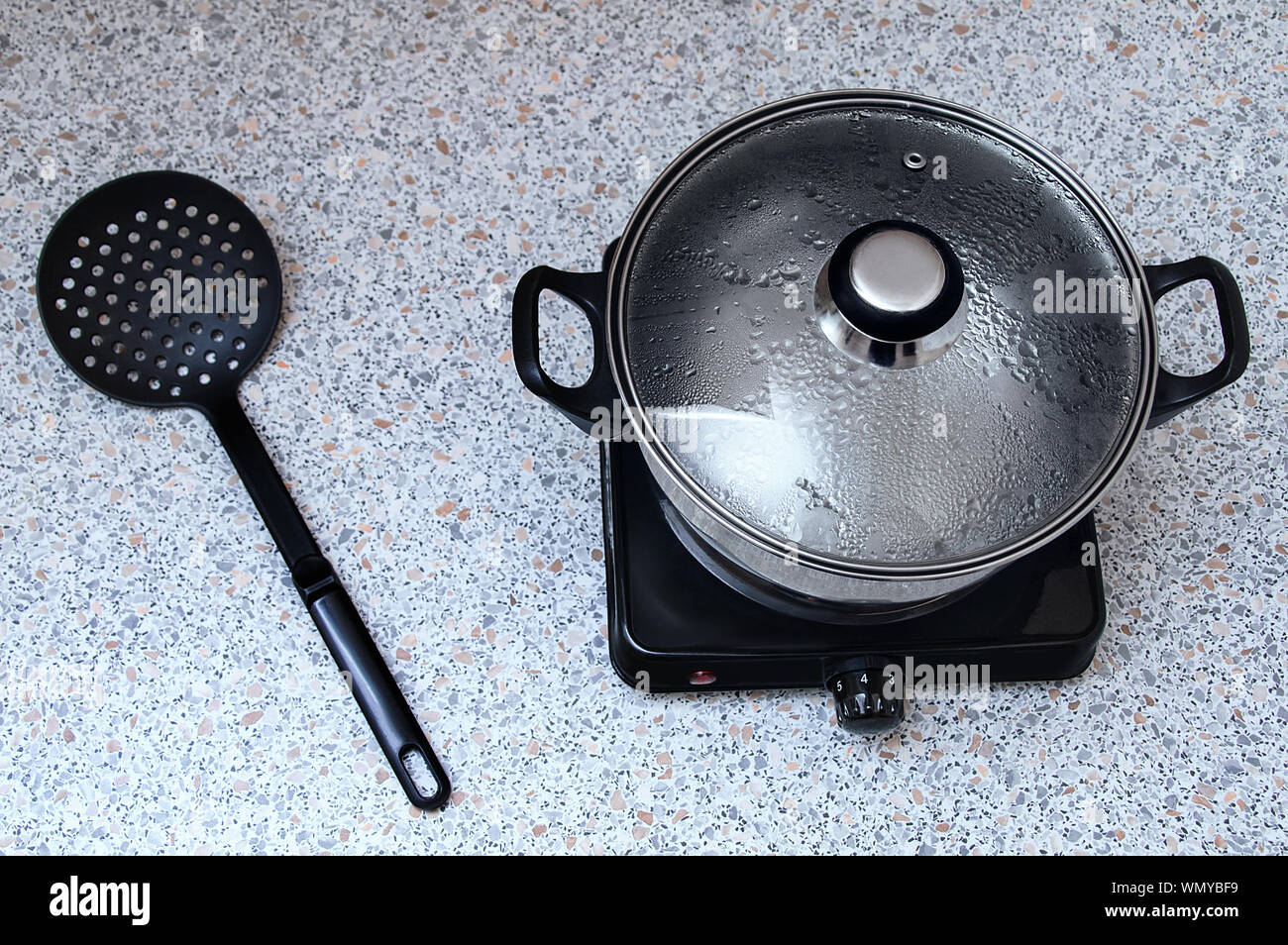 Pentola su una piccola stufa elettrica con un coperchio per un paio. Sul tavolo è un nero in plastica cucchiaio skimmer. Vista da sopra. Foto Stock