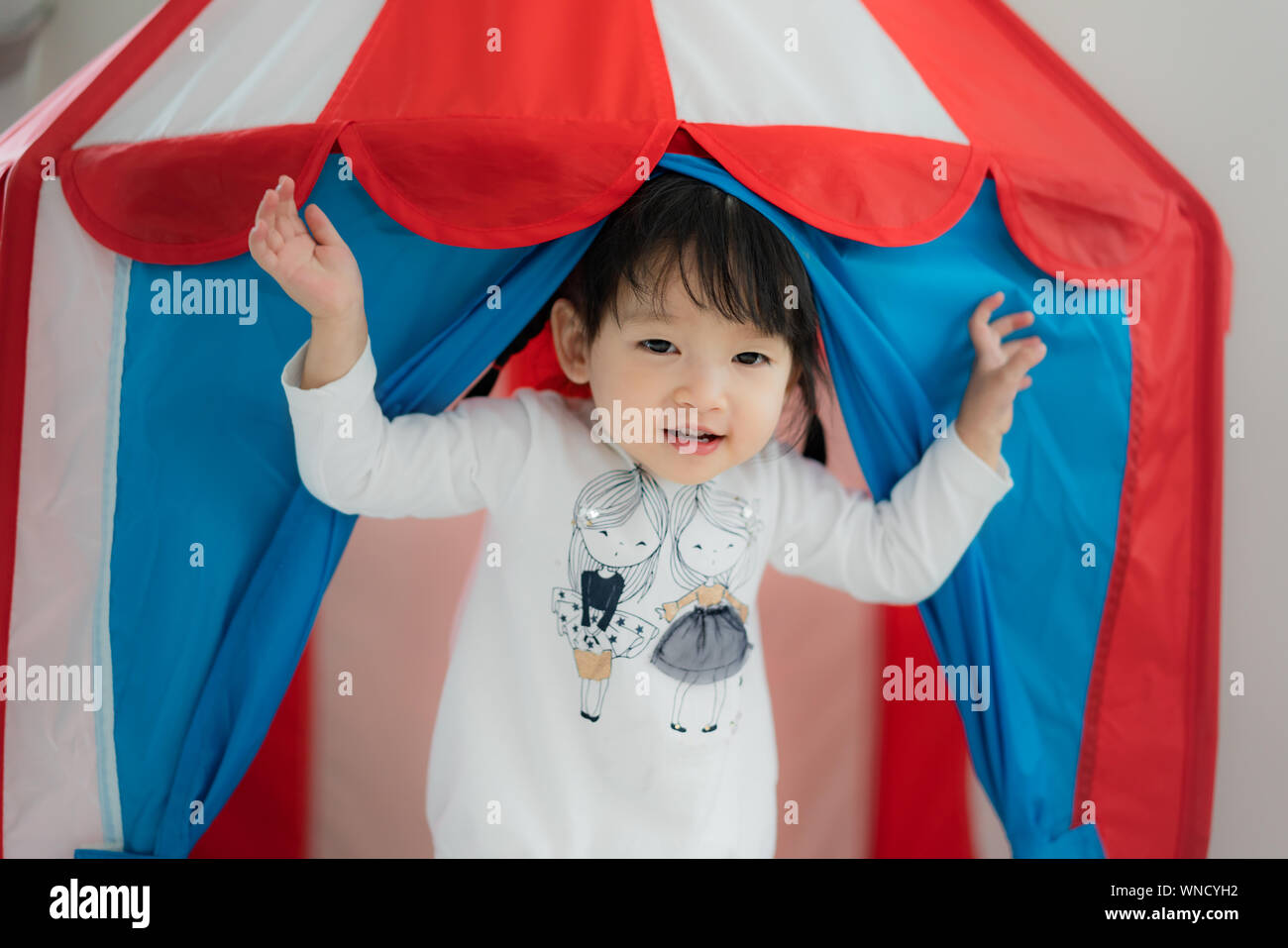 Asian toddler carina ragazza in piedi in abito bianco in tenda lodge e sorriso nel salotto di casa. Foto Stock