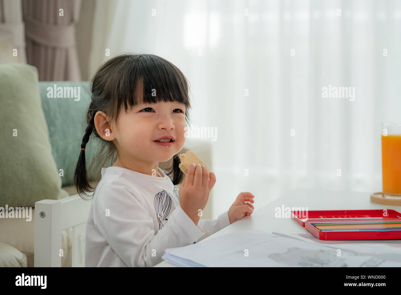 Felice toddler asiatica ragazza con un grande sorriso mentre mangia wafer quando ella disegno in soggiorno a casa sua. Foto Stock