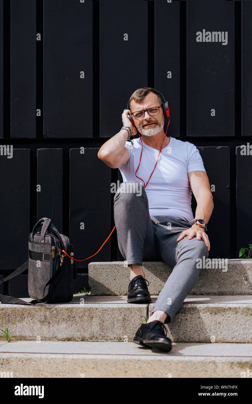 Barbuto uomo maturo seduto sui gradini con le cuffie durante i momenti di relax in una giornata di sole Foto Stock