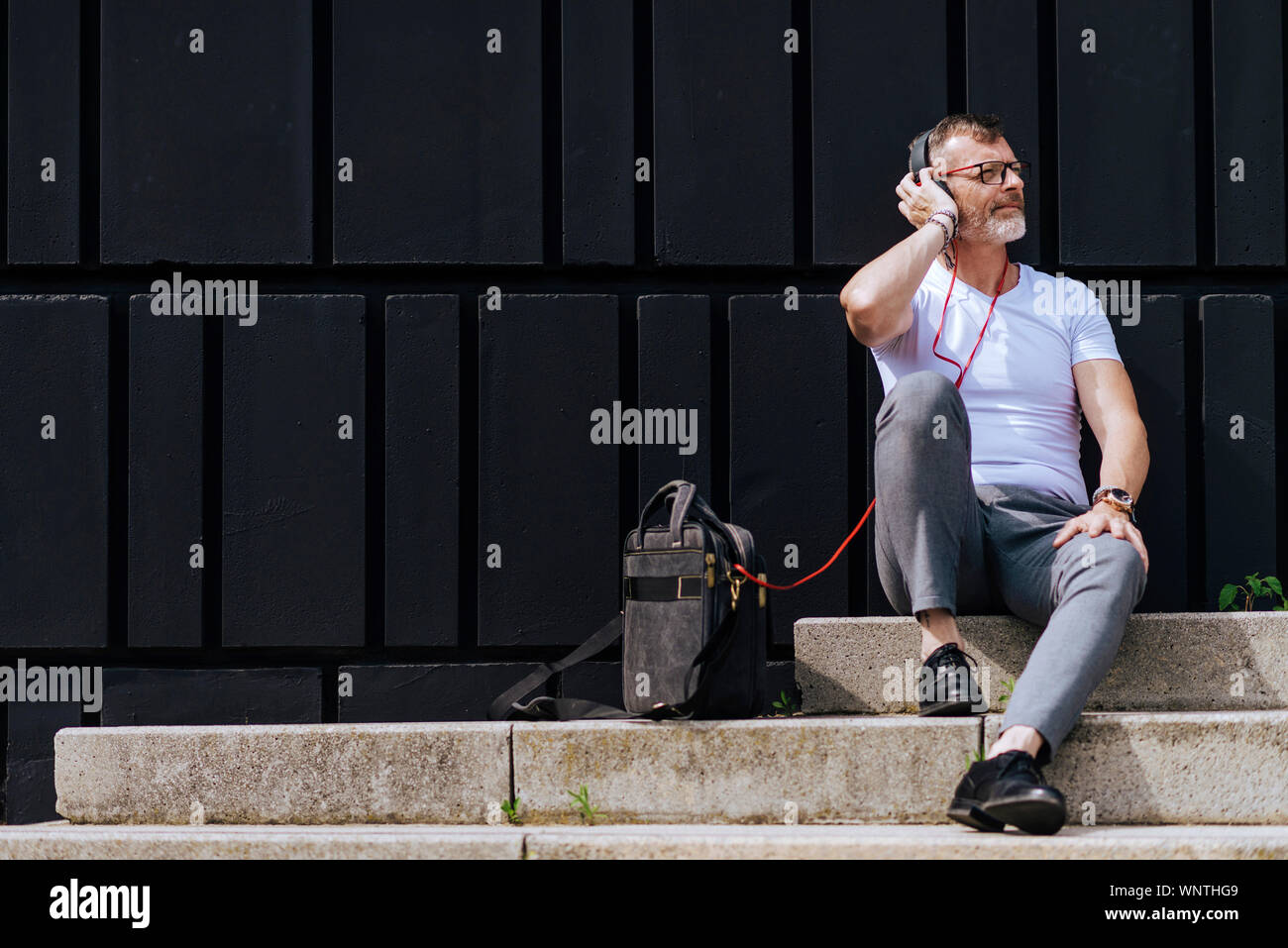 Barbuto uomo maturo seduto sui gradini con le cuffie durante i momenti di relax in una giornata di sole Foto Stock