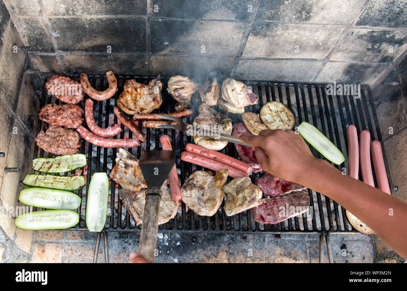 Persona assortiti di cottura la carne e le verdure su un barbecue all'aperto utilizzando utensili di metallo in un elevato angolo di prima persona POV cercando don al grill Foto Stock