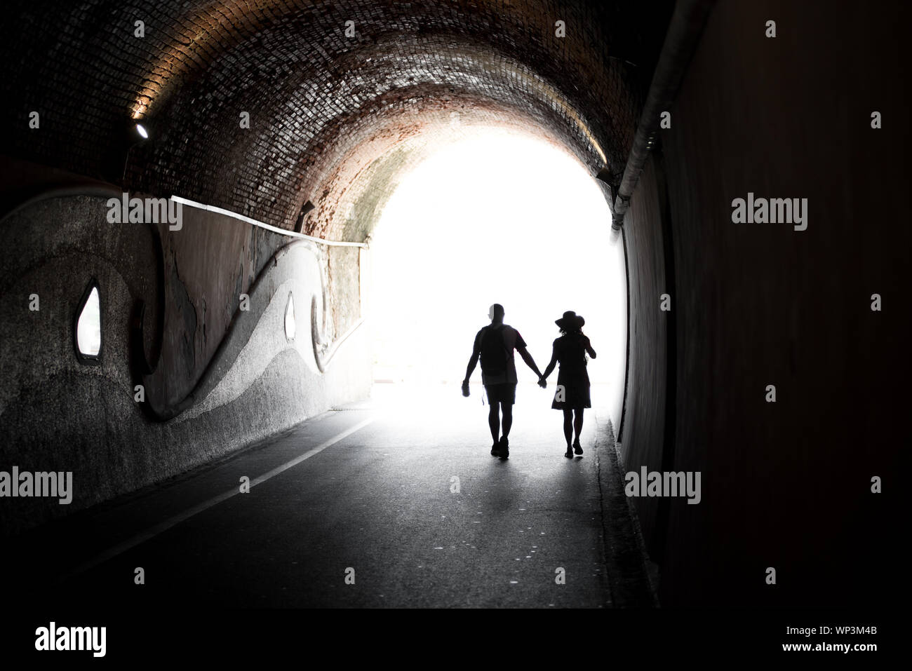 Giovane camminando mano nella mano attraverso una coperta tunnel ad arco o sottopassaggio stagliano contro la luce brillante alla fine in una immagine concettuale Foto Stock