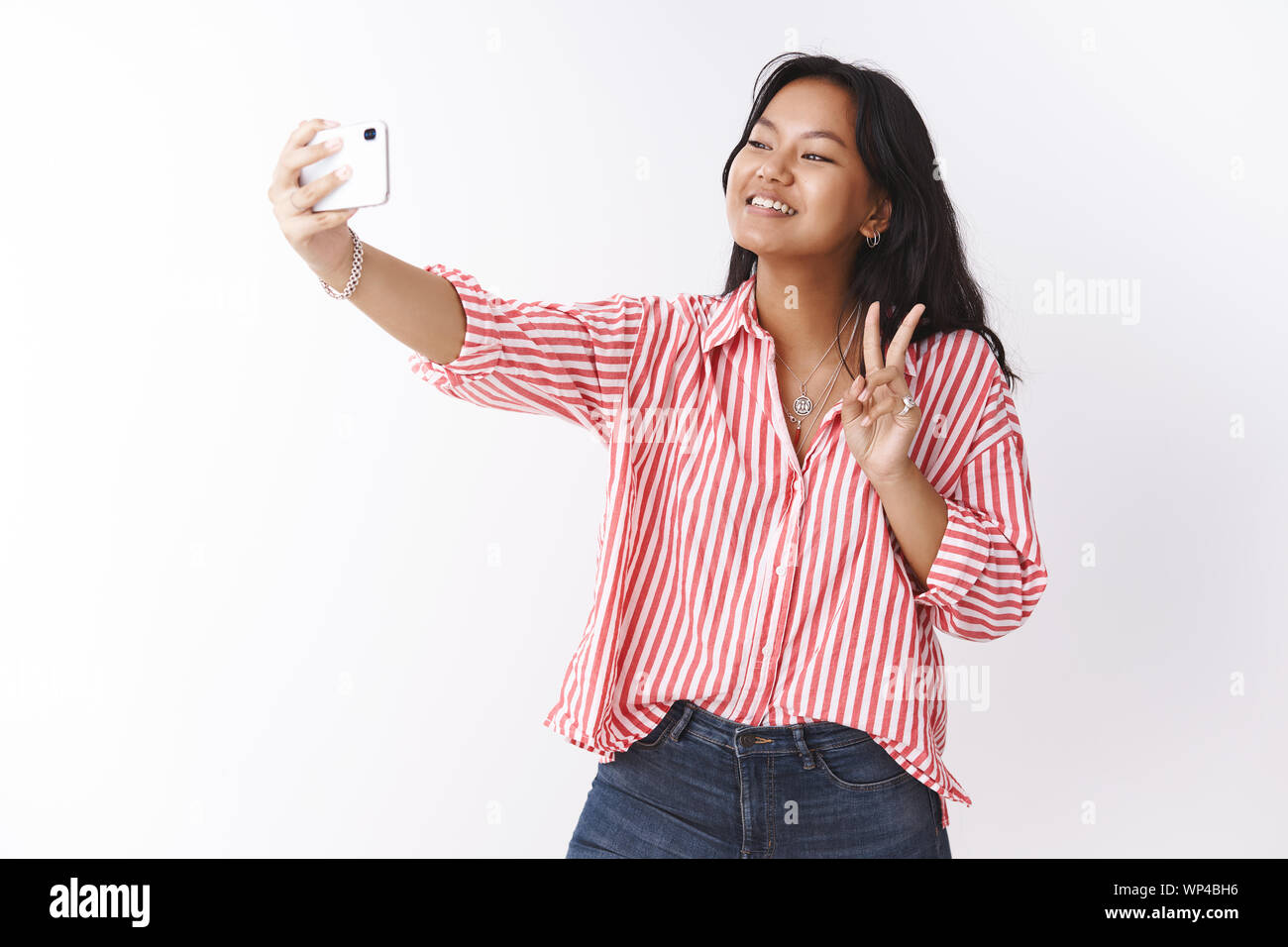 Ragazza carina tenendo selfie sul nuovo smartphone. Ritratto di uscenti e socievole giovane femmina comunicativa nella camicia a righe che mostra segno di vittoria foto Foto Stock