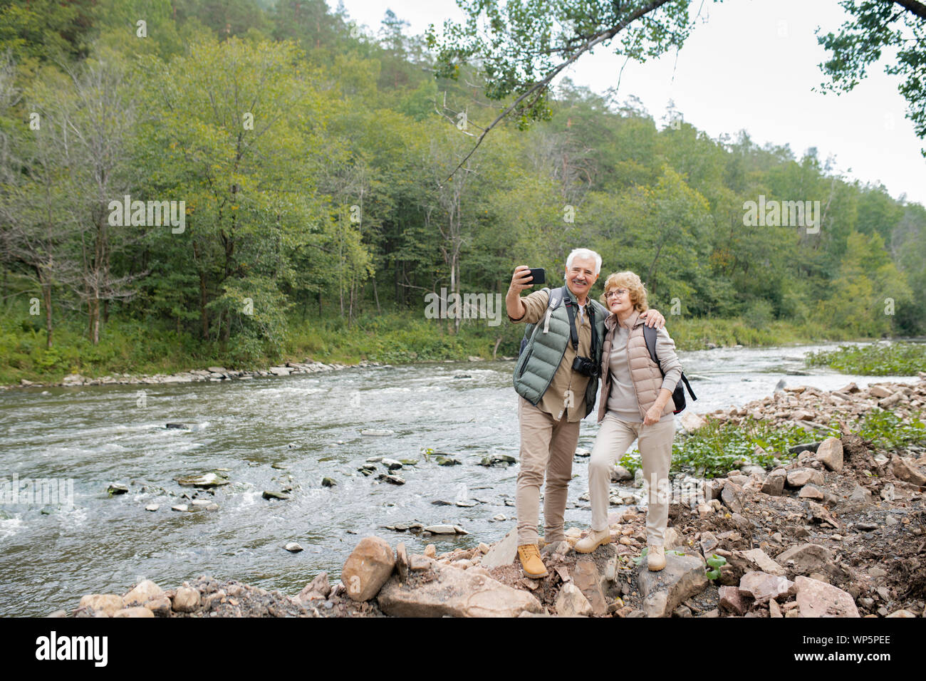 Due allegri escursionisti maturo con zaini rendendo selfie sulla banca del fiume Foto Stock