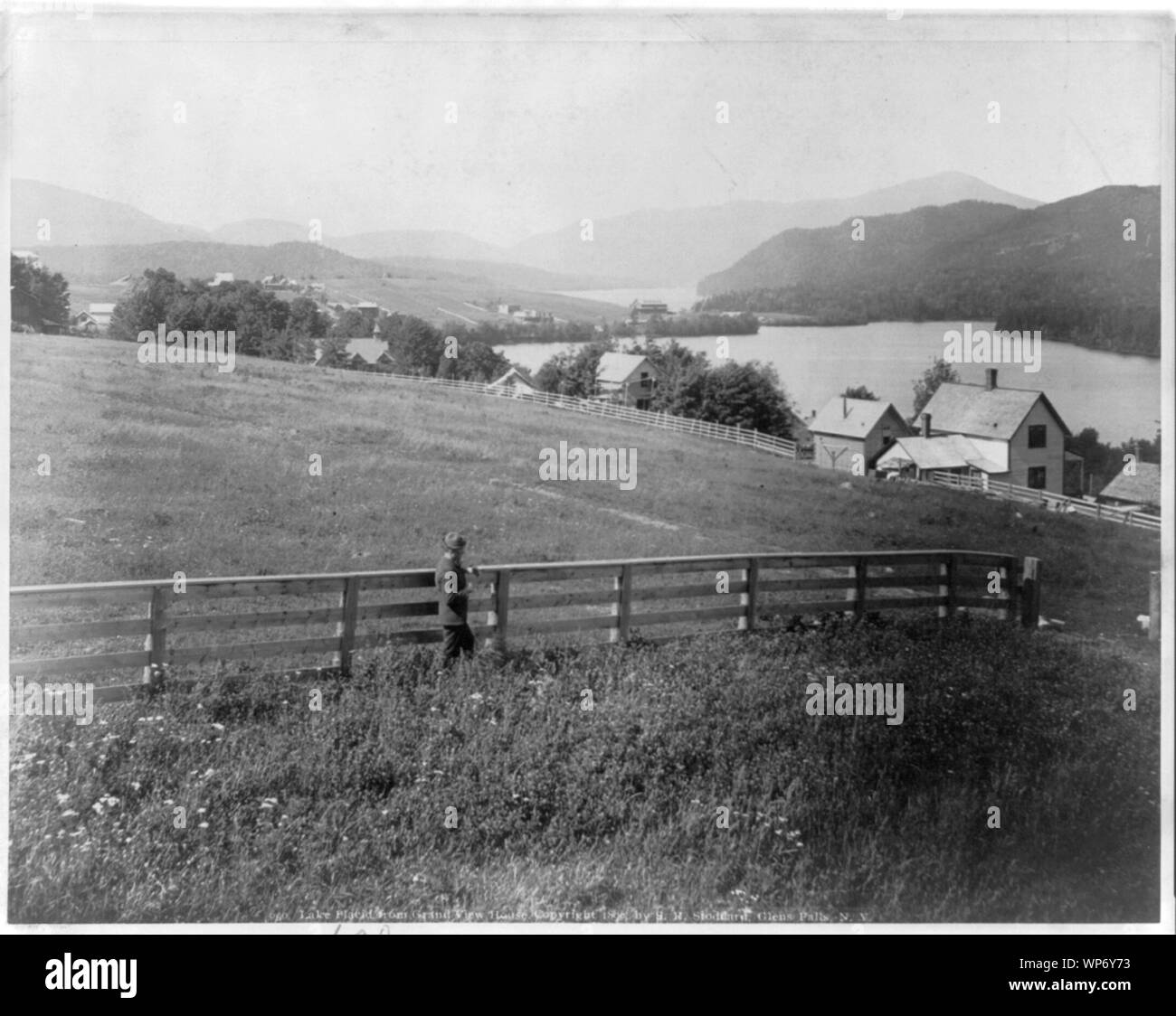 Lake Placid dal Grand View House, [Aidrondack montagne, New York, con un uomo in piedi da recinzione in campo in primo piano] Foto Stock