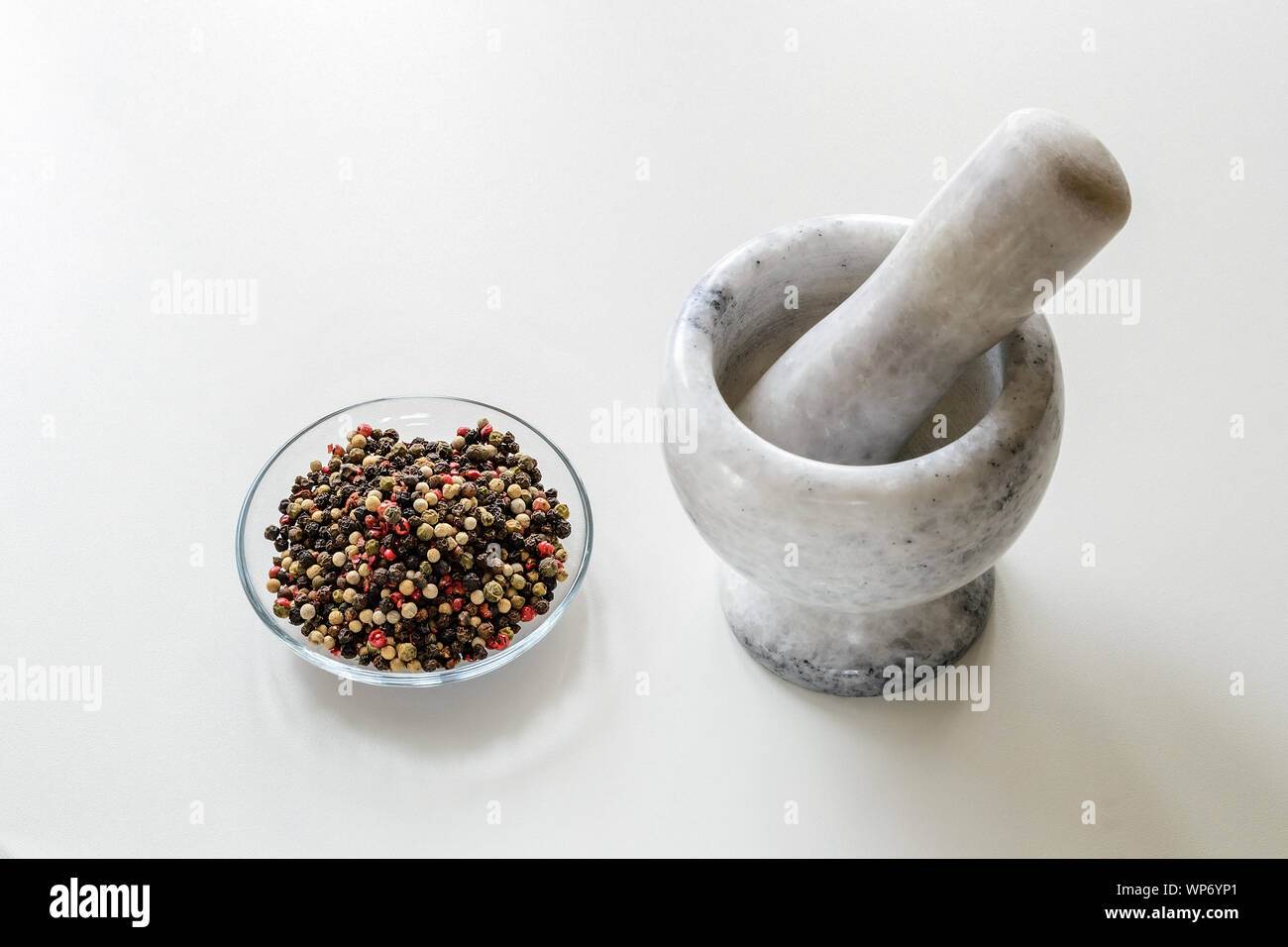 Piattino in vetro con un mix di grani di pepe e di marmo o granito mortaio con pestello per il manuale di macinazione di spezie su una cucina bianca tabella. Moderni utensili di pietra Foto Stock