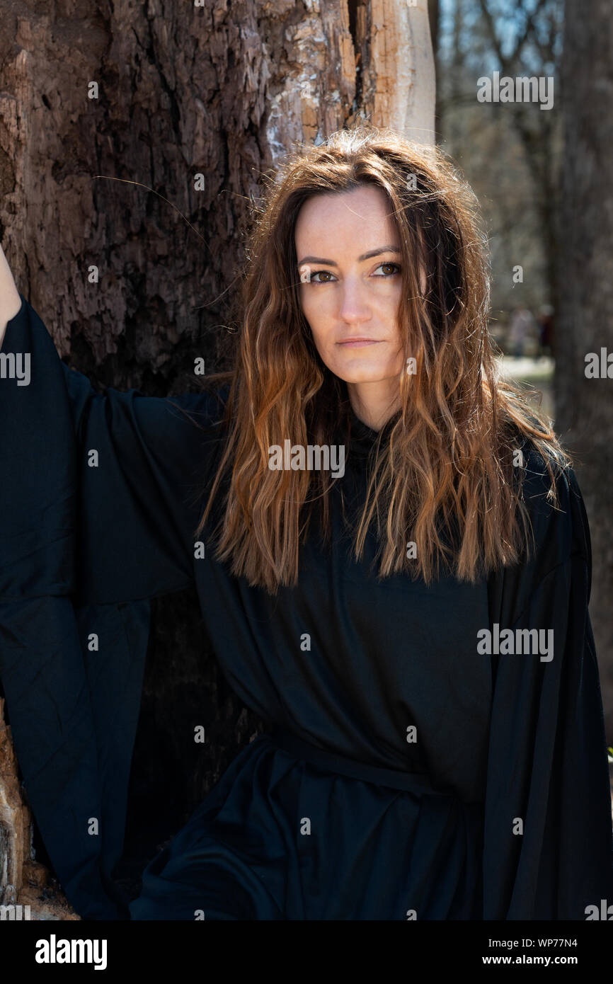 Giovani Tall Woman con scuri capelli lunghi in black robes nel profondo della foresta. Streghe. Halloween e concezione gotica. La stregoneria e magia. Foto Stock