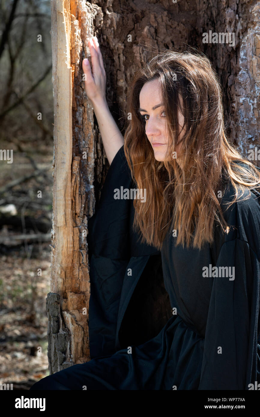 Giovani Tall Woman con scuri capelli lunghi in black robes nel profondo della foresta. Streghe. Halloween e concezione gotica. La stregoneria e magia. Foto Stock