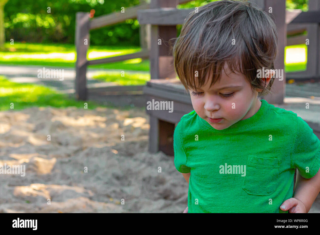 Un bambino in una camicia verde, a circa due anni di età, sorge nella sabbia di un parco giochi ma sembra essere nel profondo del pensiero. Foto Stock