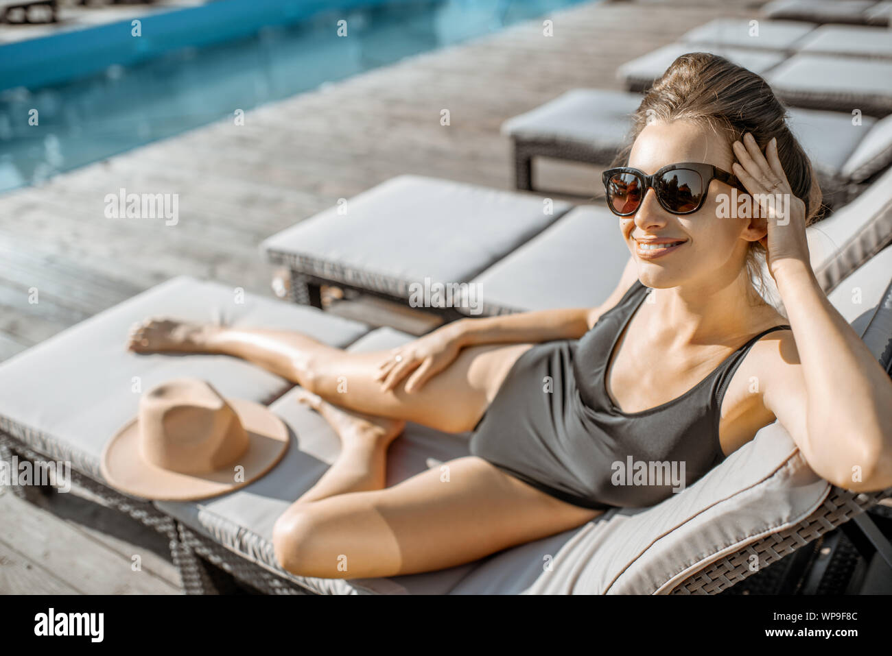 Donna in costume nero sdraiato sul lettino vicino alla piscina all'aperto. prendere il sole su un tempo di estate Foto Stock